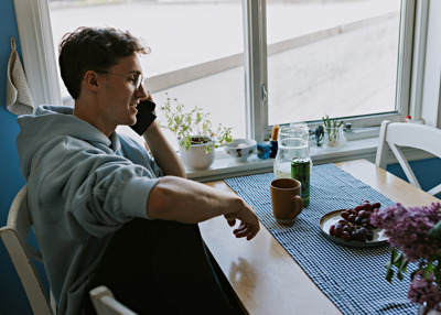 Mann sitter ved et kjøkkenbord og snakker i telefonen. På bordet står en kaffekopp og et fat med druer. 