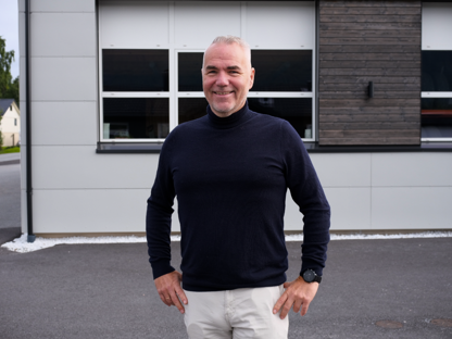 Smilende mann i svart genser står foran en moderne bygning. Utebilde med vinduer og mørk fasade.