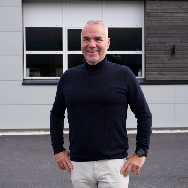 Smilende mann i svart genser står foran en moderne bygning. Utebilde med vinduer og mørk fasade.