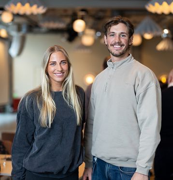 Kvinne med langt lyst hår og mann med kort hår, begge kledd i collegegenser, står sammen og smiler mot kamera.