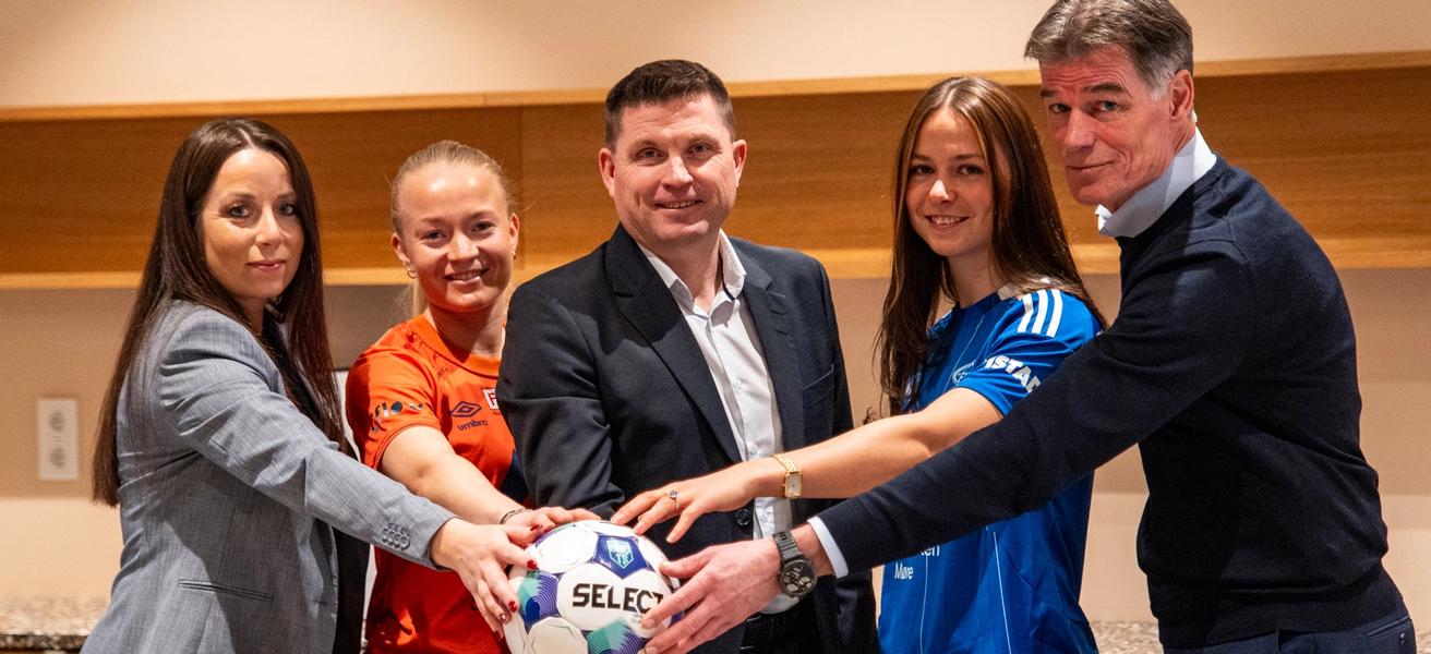 Tre kvinner, to med fotballdrakt, og to menn står sammen og holder en hånd hver på en fotball.
