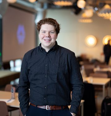 Ung mann med mørk skjorte, hendene i lommen. Smiler til kamera. Blurret bakgrunn