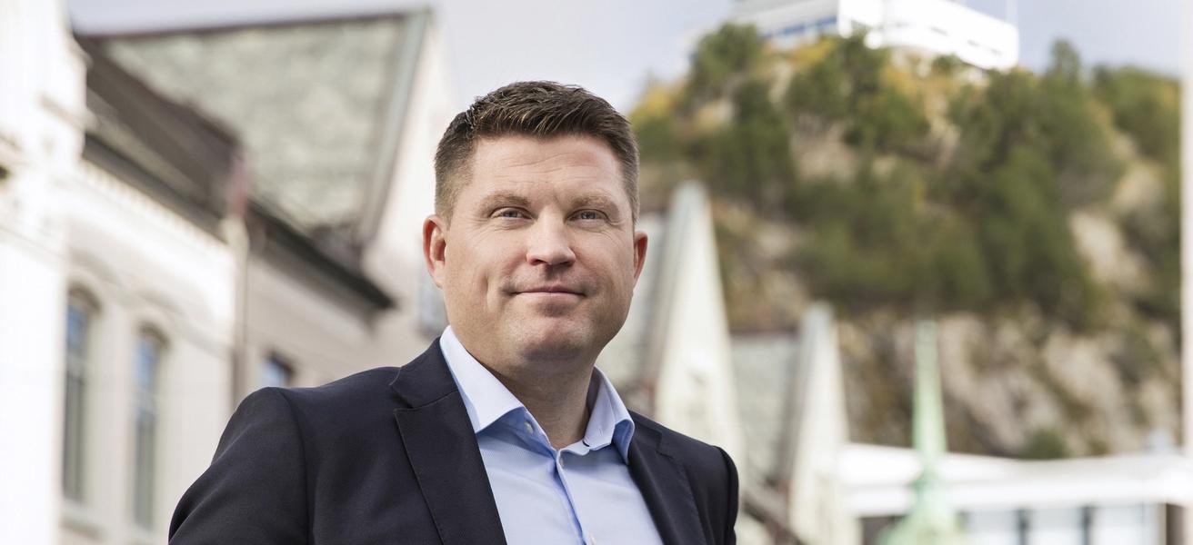 Dresskledd mann, stående med fjellstua i Ålesund bak seg.
