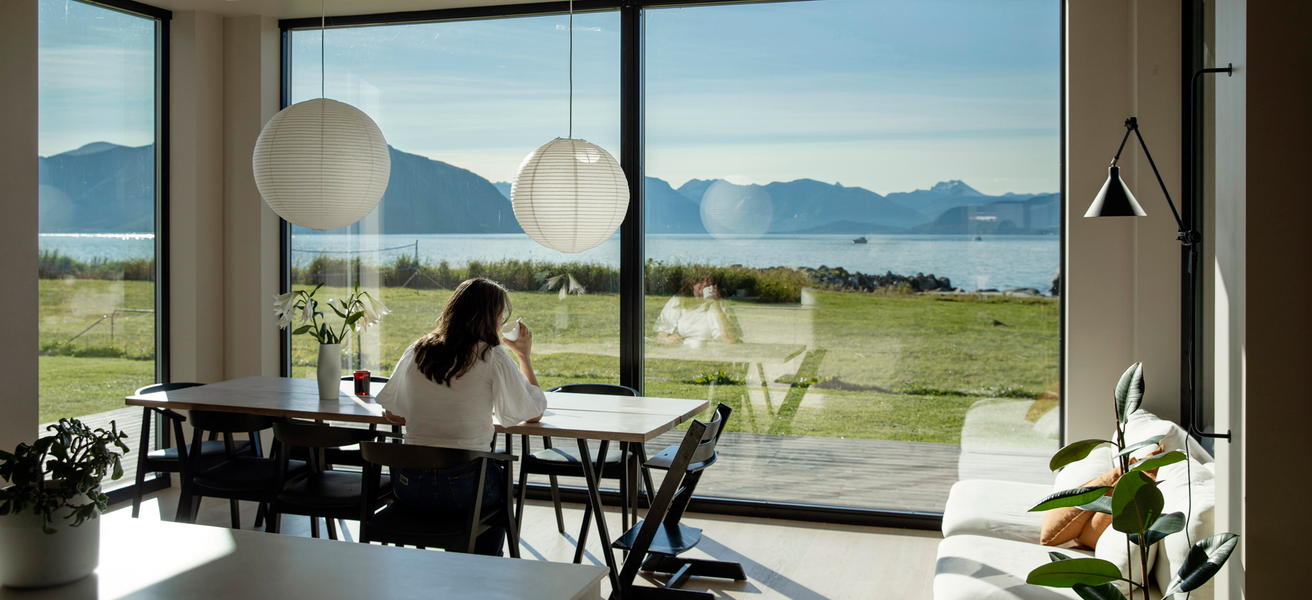 Kvinne sitter ved kjøkkenbord, ser ut av vindu mot havet.