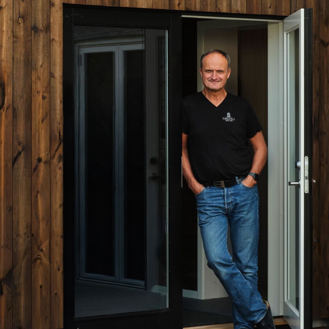 Daglig leder i Tingvoll Dør og Vindu står i en spesialtilpasset ytterdør ved bygg med trekledning, produsent av skreddersydde dører og vinduer.