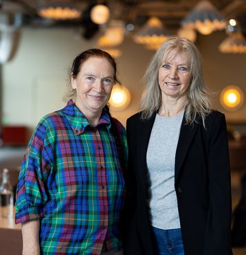 To kvinner som står sammen og ser i kamera. En kledd i t-skjorte og dressjakke, den andre i rutete skjorte.
