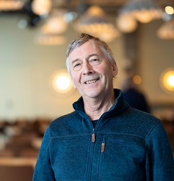 Mann med grått hår, blå fleecejakke. Smiler og ser i kamera. Blurret bakgrunn.