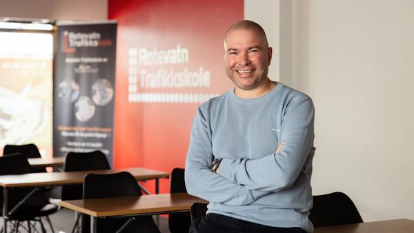 Mann med lys genser sitter på en undervisningspult, smiler til kamera. Rotevatn Trafikkskole står på en rollup i bakgrunn.