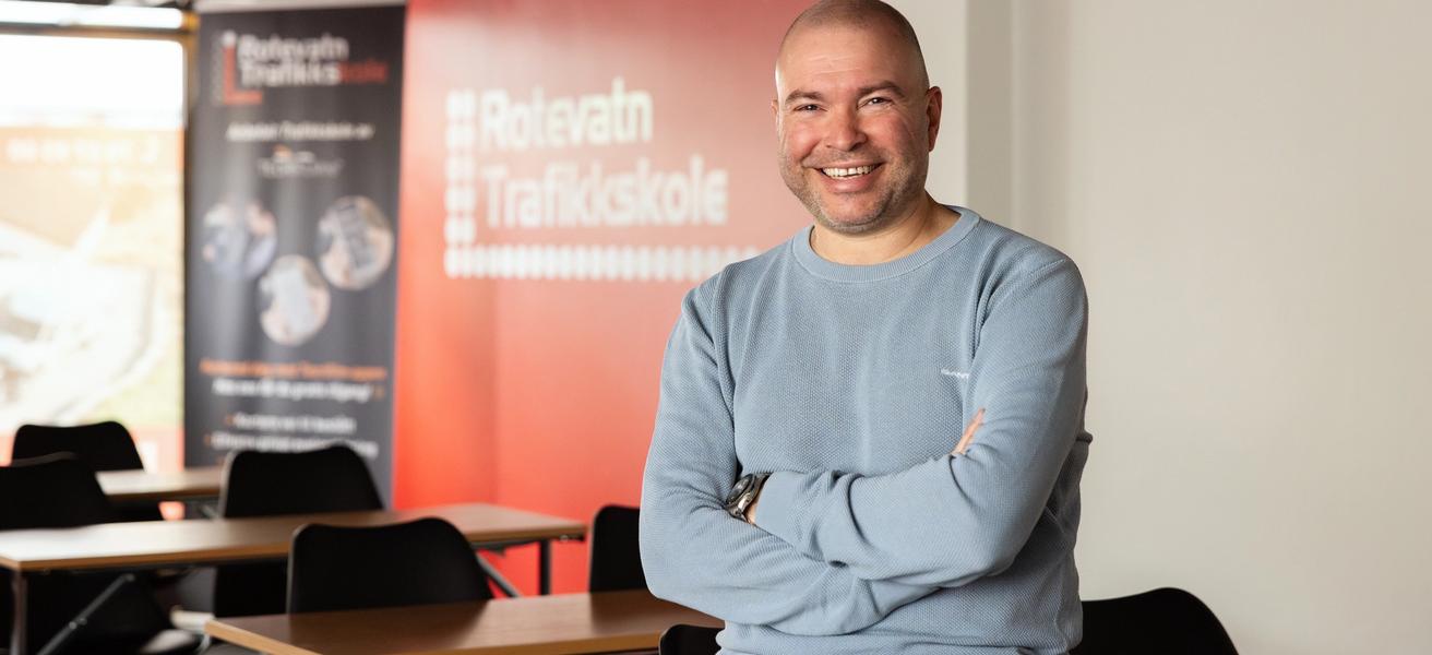 Mann med lys genser sitter på en undervisningspult, smiler til kamera. Rotevatn Trafikkskole står på en rollup i bakgrunn.