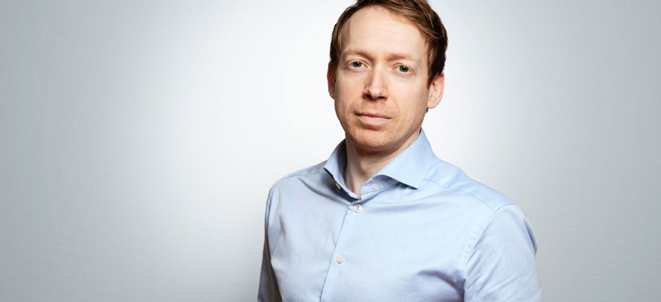 Portrett av Christian Balsnes i lyseblå skjorte mot en lys grå bakgrunn. Han har et rolig uttrykk og ser direkte mot kamera.