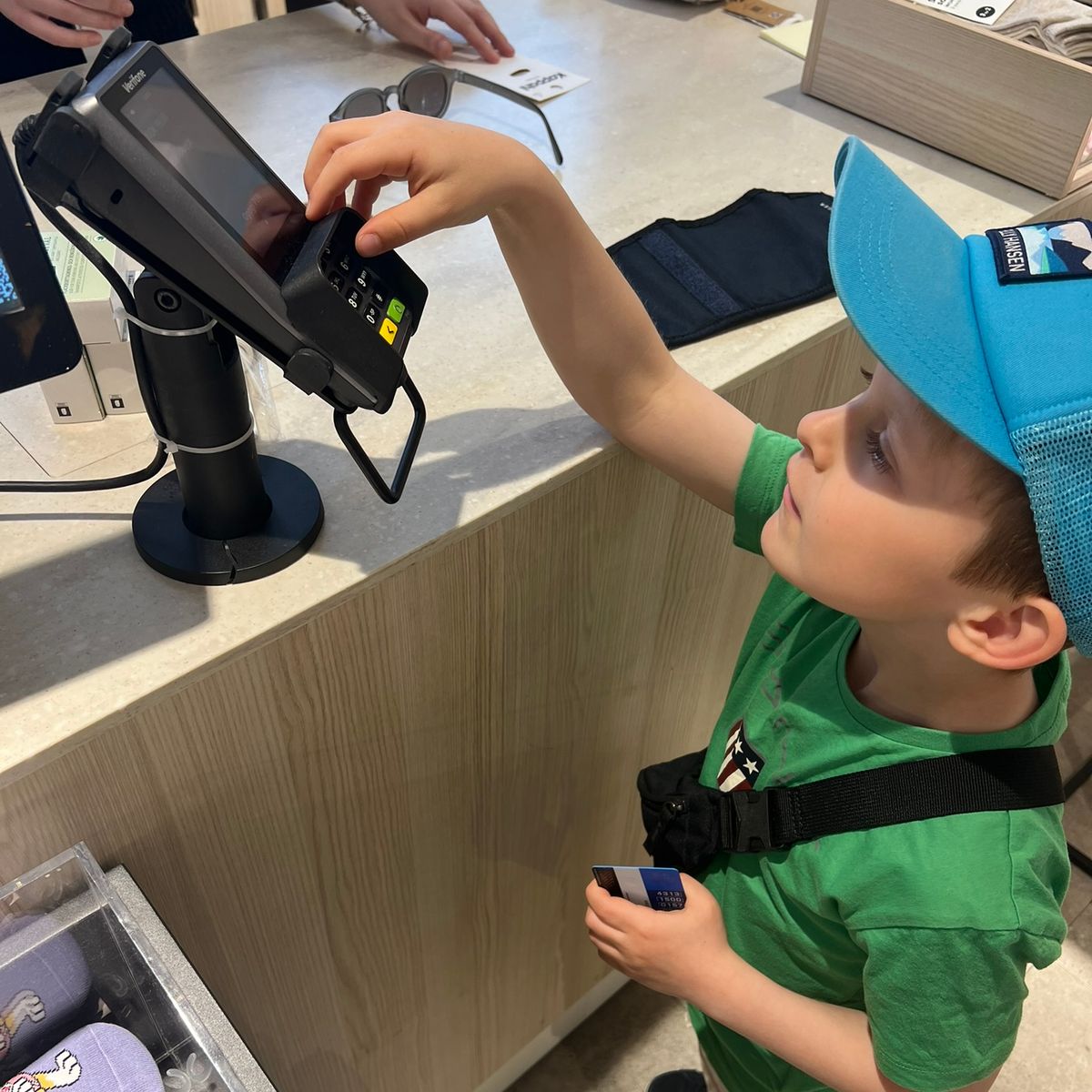 Liten gutt med fargerik t-skjorte og caps som betaler for en vare i en butikk med bankkort.