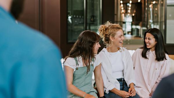 Tre studenter, jenter, sitter sommen, vårlig kledd. Ser på hverandre og prater, og ler.
