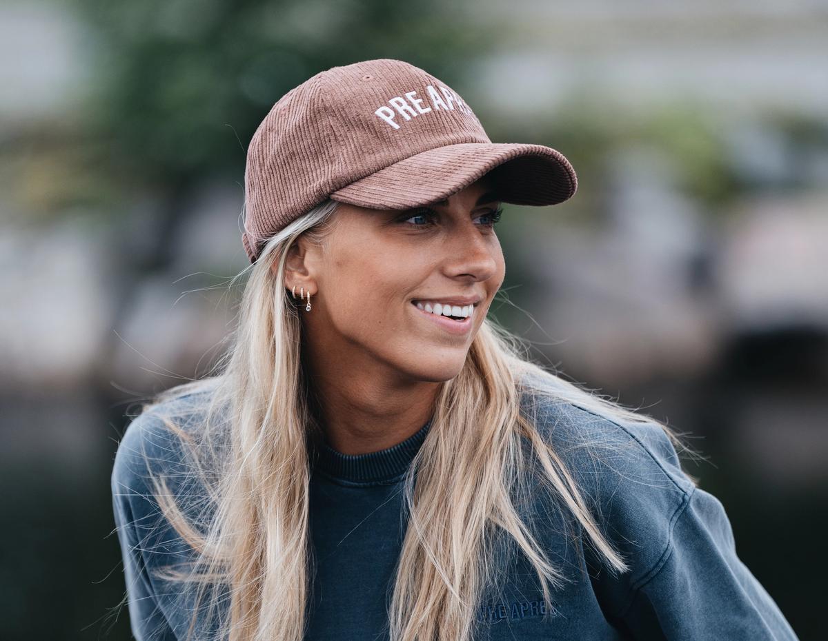 Kvinne med langt lyst hår og caps. Smiler og ser på noen utenfor bildet. Lener seg lett fremover og har hendene i lommen.