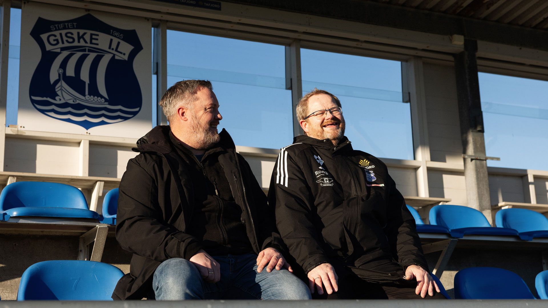 To menn sitter på en fotballtribune. De smiler. 