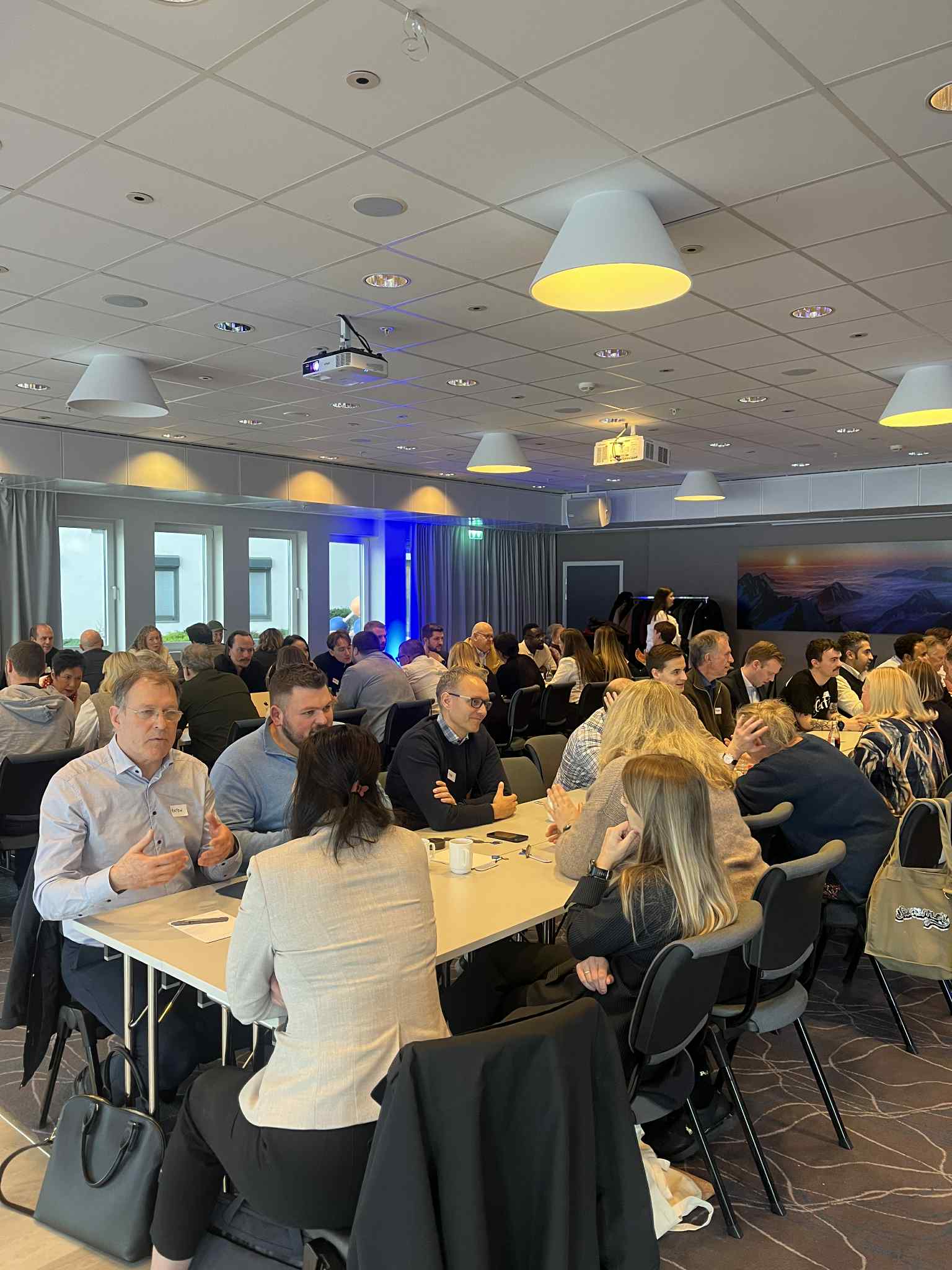 Forsamling med mennesker som sitter på langbord i et kurslokale - i samtale med hverandre