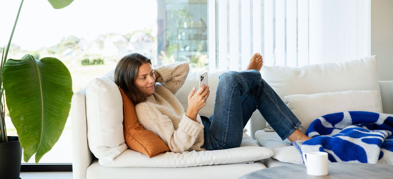 Kvinne sitter i sofa, ser på mobil hun holder i hånden.