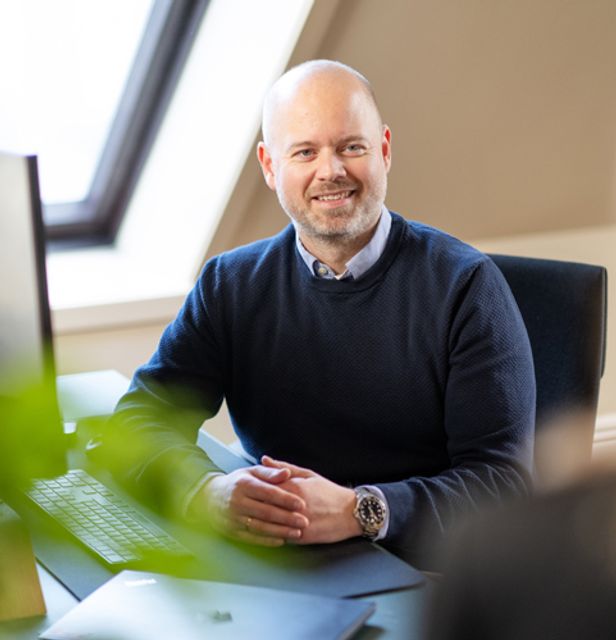 Mann som sitter med foldet hender på en pult med PC og tastatur og snakker med en person vi bare ser bakhodet på