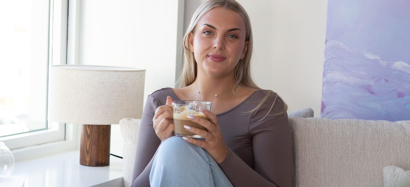 Ung kvinne i et moderne innemiljø, i sofa med en kopp kaffi. Smiler til kamera.