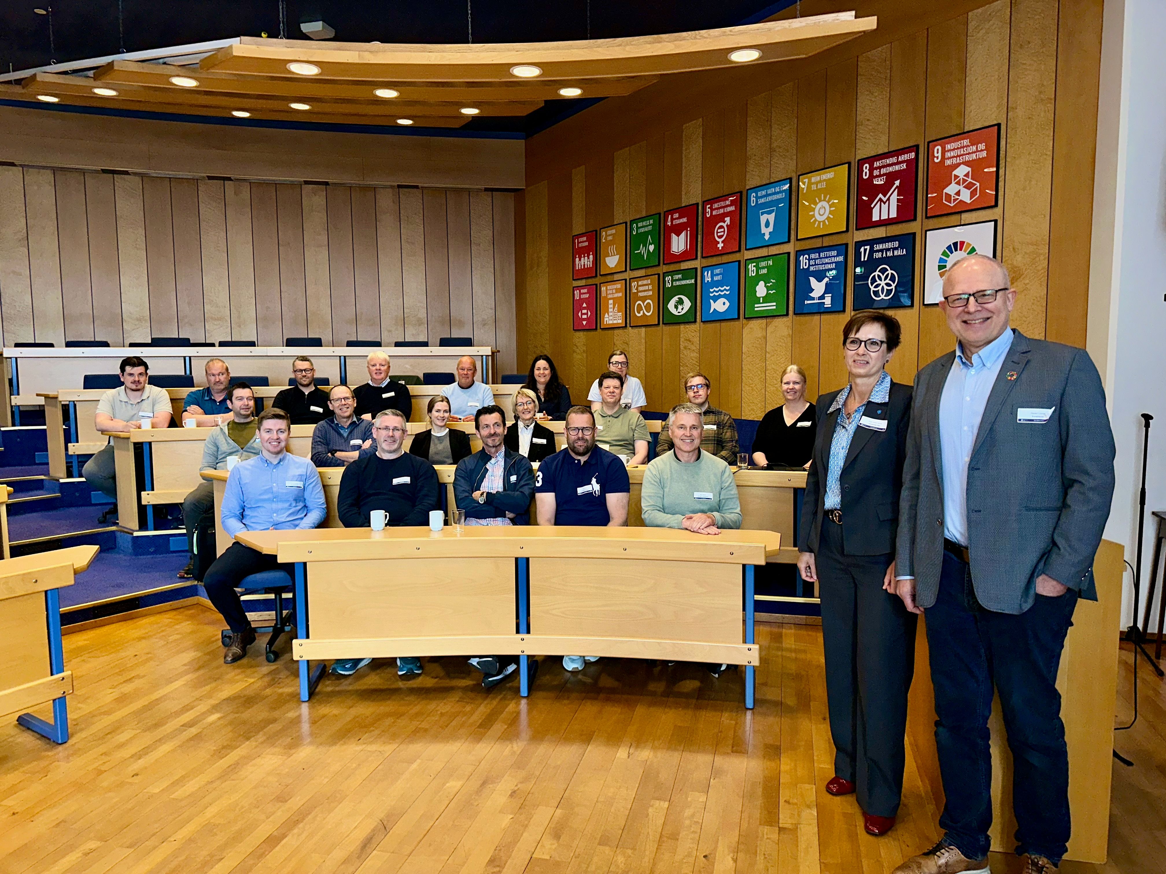 Gruppebilde fra samling i et auditorium: deltakere sitter ved benker, to personer står foran, og på veggen henger ikoner for FNs bærekraftsmål. 