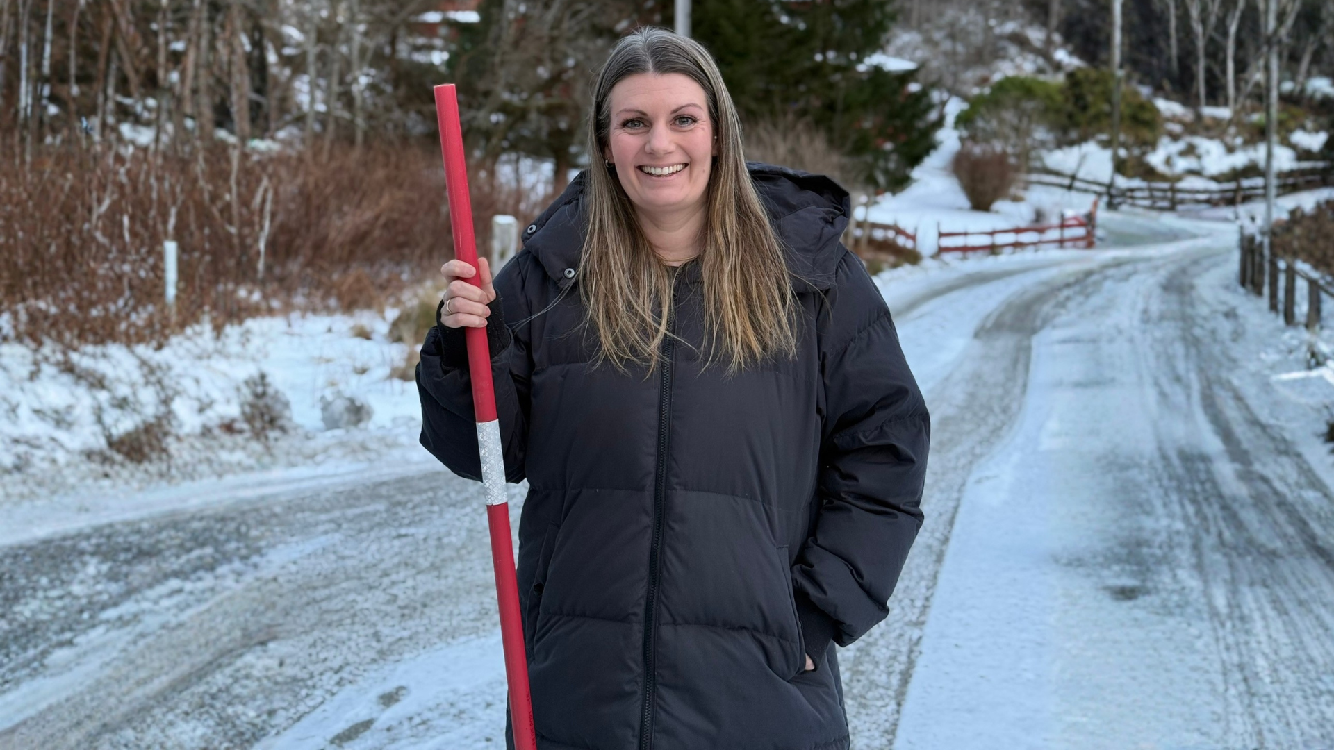 Elisabeth Pedersen Nerdal står utendørs på en vintervei og holder en rød brøytestikke. Hun er kledd i mørk vinterjakke, og veien er dekket av snø og is.