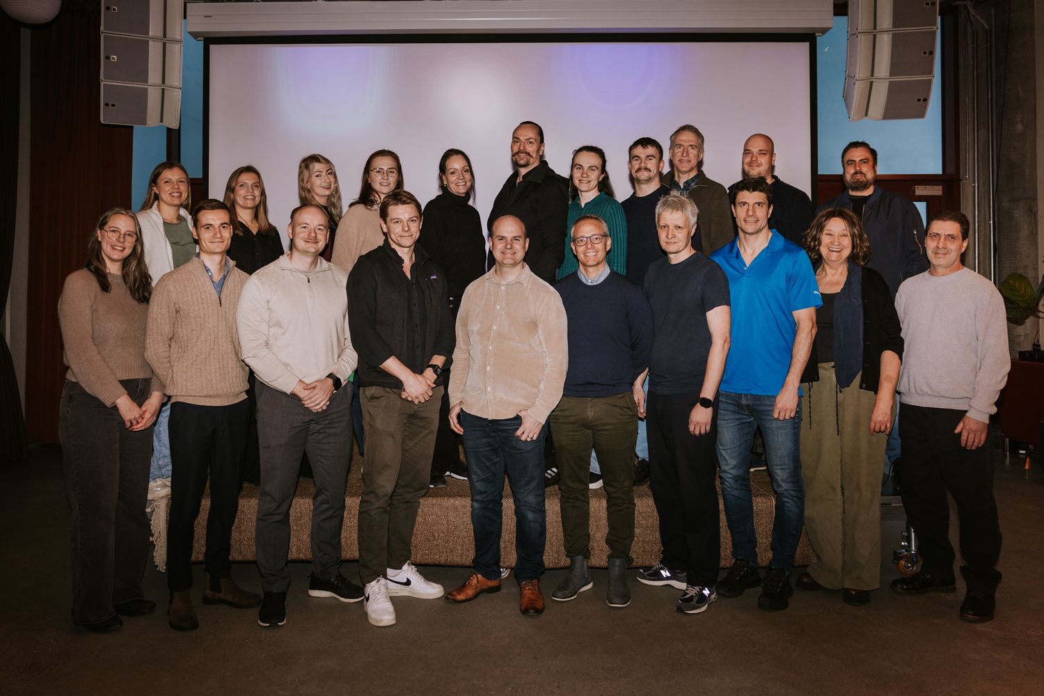 To oppstilte rekker med mennesker, kvinner og menn, står foran et lerret i et arrangementslokale.