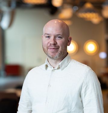 Mann med kvit skjorte, smiler til kamera - blurret bakgrunn.