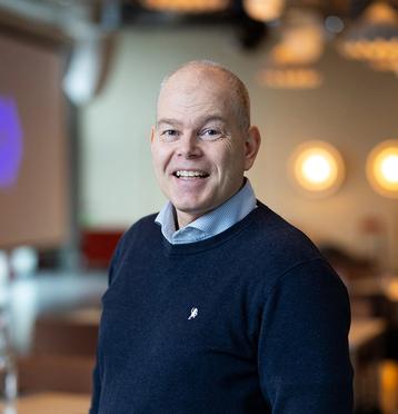 Mann med mørk blå genser og skjorte, smiler og ser i kamera. Blurret bakgrunn.