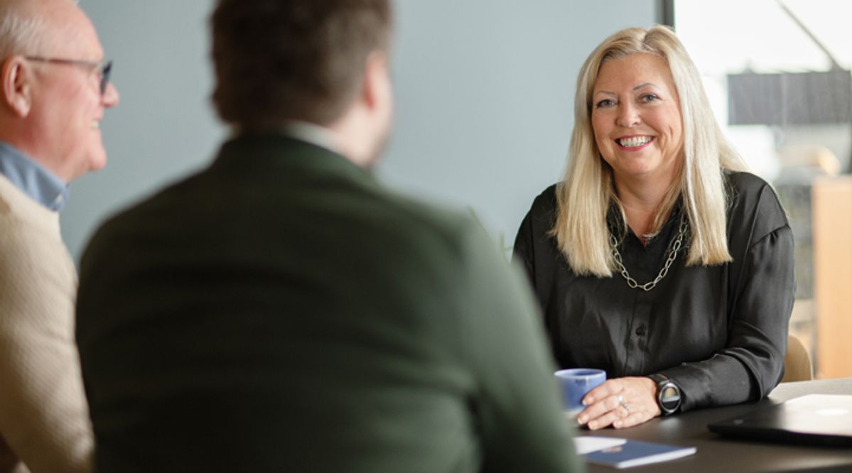 Tre personer i et profesjonelt møte, hvor en smilende kvinne i svart skjorte og blondt hår holder en kopp og deltar aktivt i samtalen. Bildet formidler samarbeid, kommunikasjon og et positivt arbeidsmiljø i en moderne kontorsetting