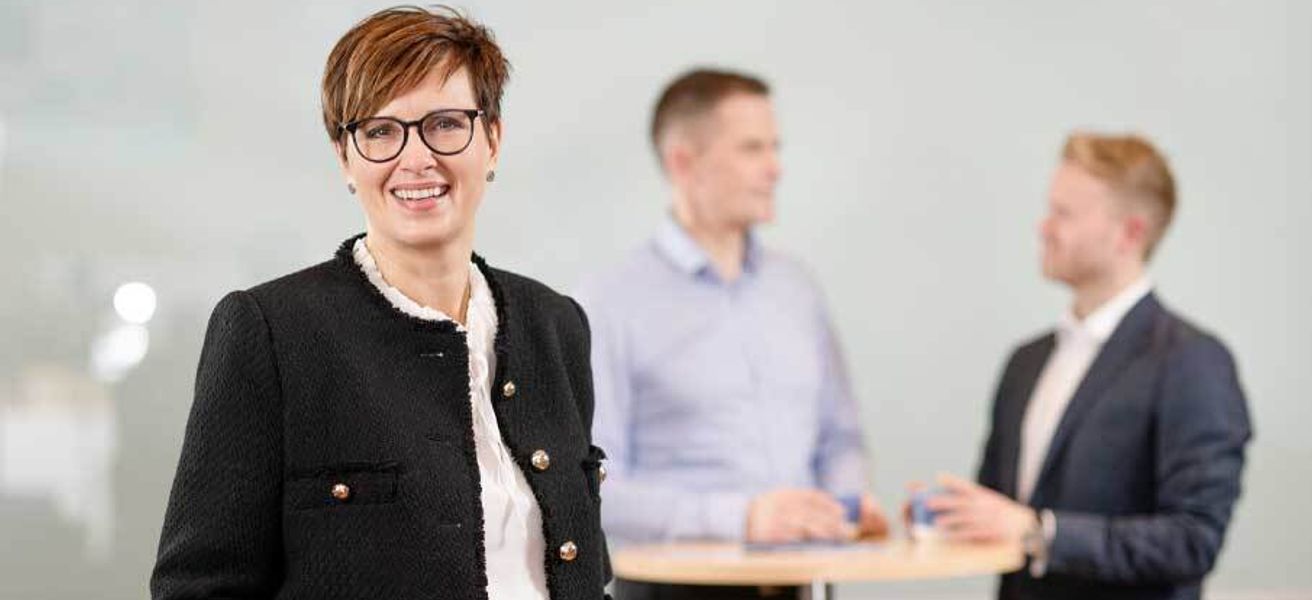 Smilende kvinne i sort jakke og briller står i fokus, mens to menn i dress og skjorte snakker ved et ståbord i bakgrunnen.