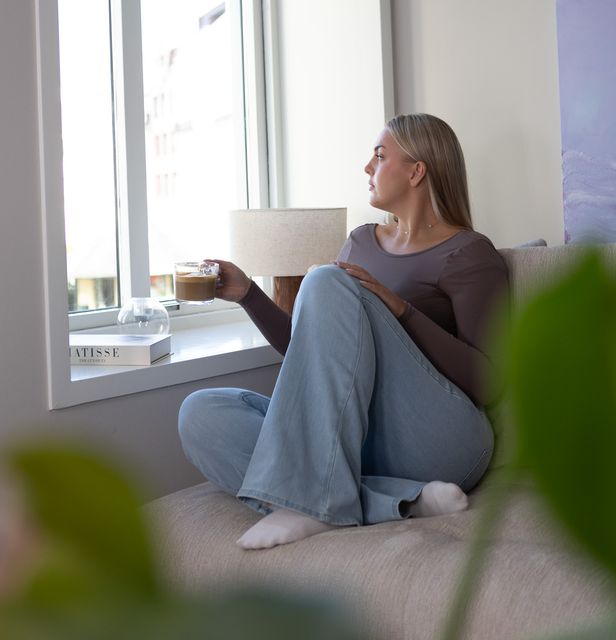 Ung kvinne som er ut av vinduet med kaffi i hånden, moderne leilighet i by.