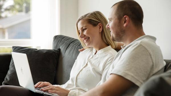 Mann og dame i lyse klær sitter i en mørk sofa med laptop på fanget. De ser på skjermen sammen.