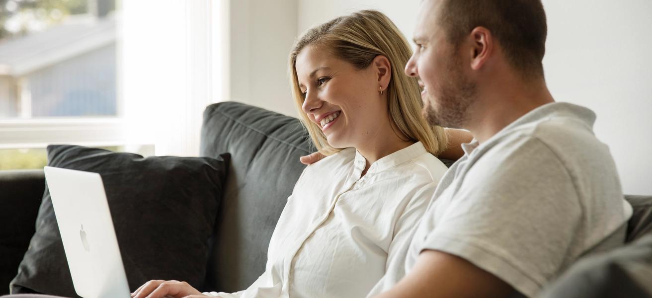Mann og dame i lyse klær sitter i en mørk sofa med laptop på fanget. De ser på skjermen sammen.