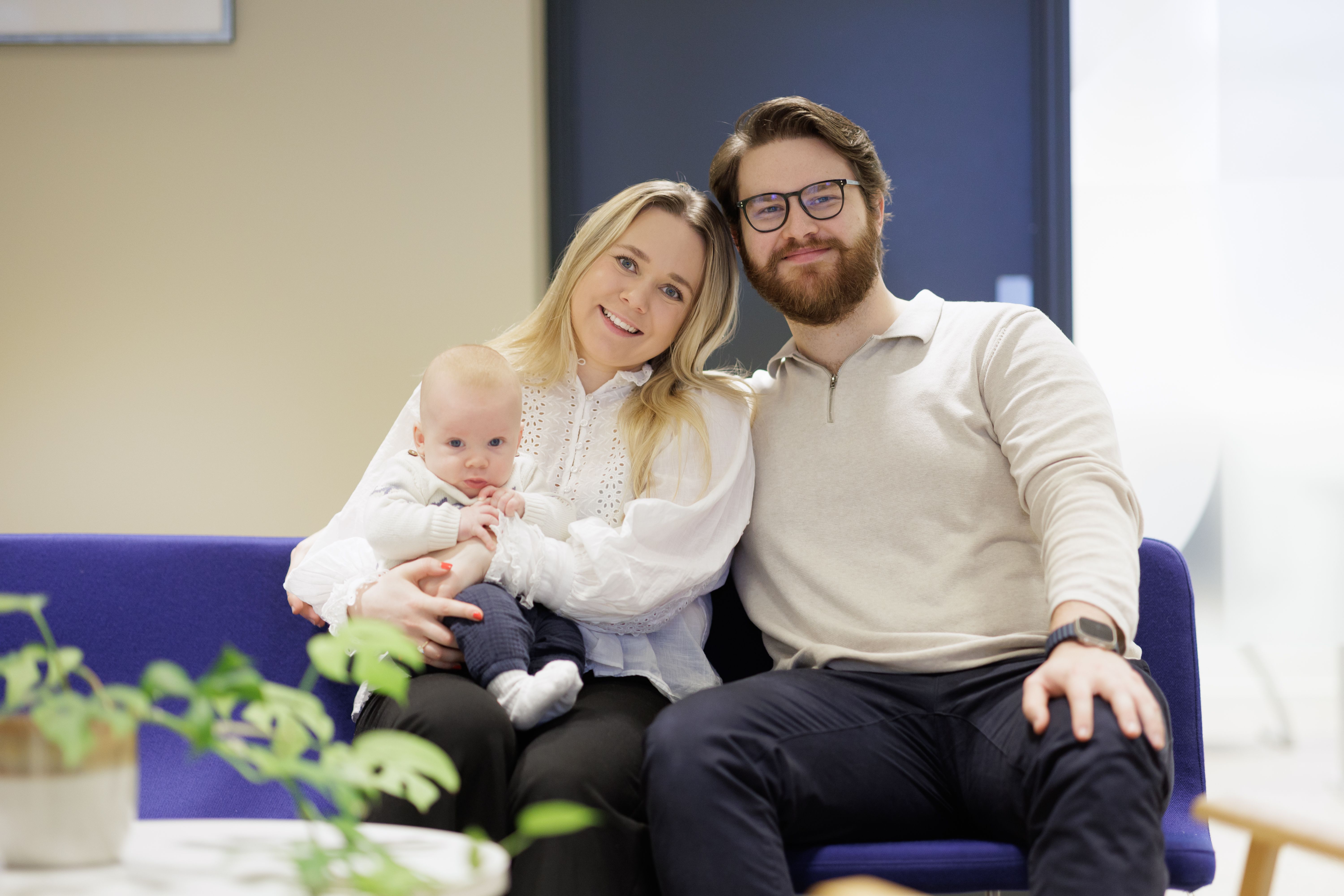 Ung par med baby på fanget, sitter på blå sofa i et kontorlokale. 