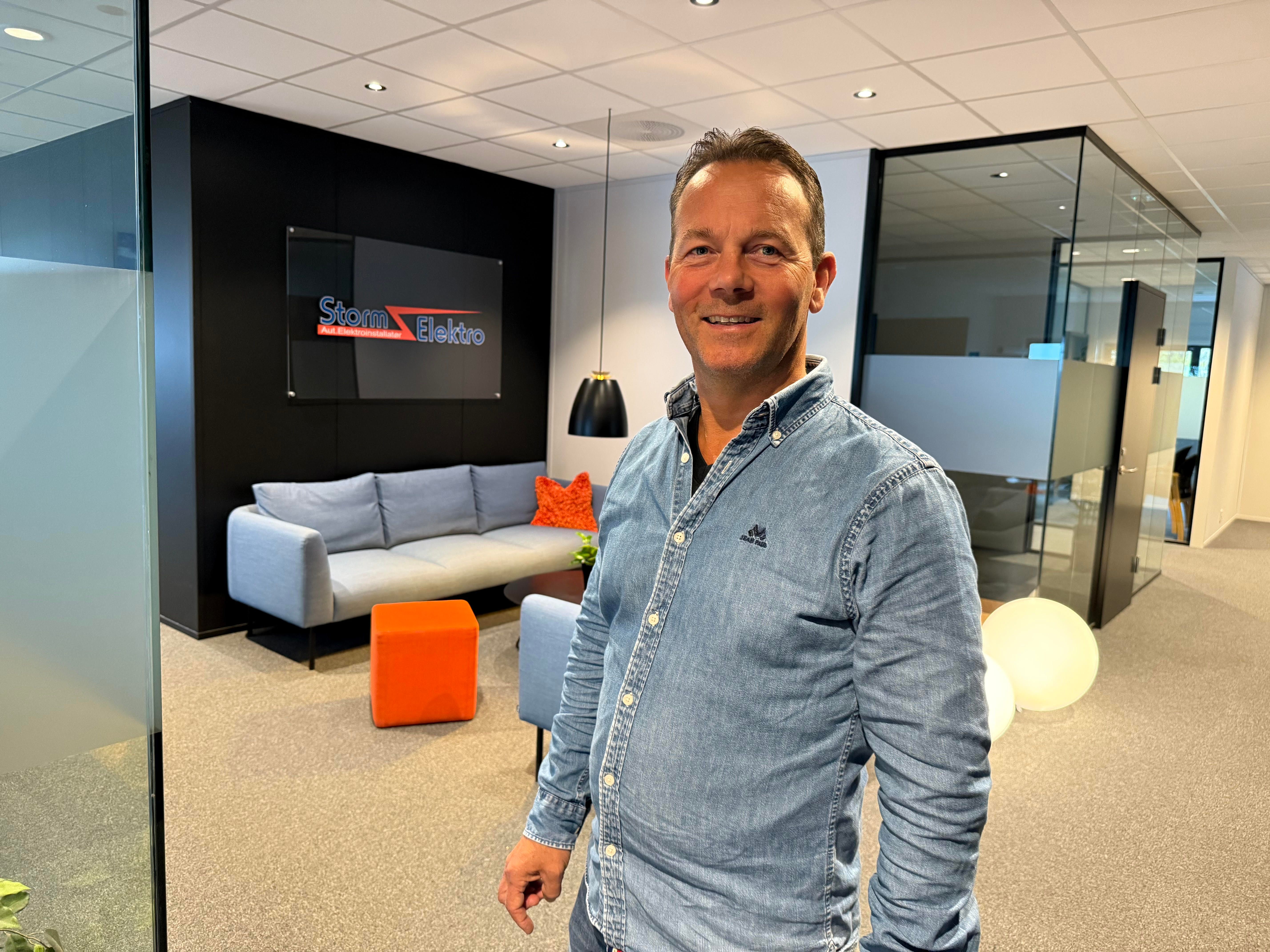 Mann med dongeriskjorte smiler mot kamera. Kontorlokaler i bakgrunn.