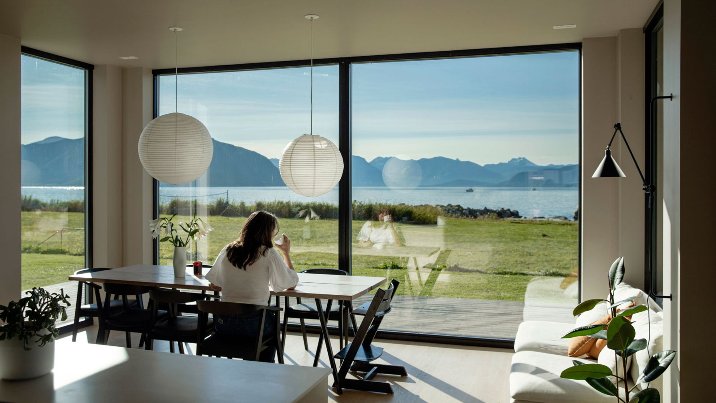 Kvinne sitter ved bord, drikker kaffe og ser ut av vindu.