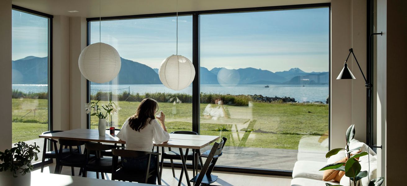 Kvinne sitter ved bord, drikker kaffe og ser ut av stort vindu med utsikt mot havet.