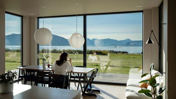 Kvinne sitter ved bord, drikker kaffe og ser ut av stort vindu med utsikt mot havet.