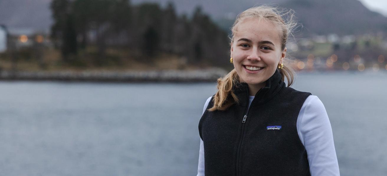 Ung jente som smiler i kamera, ute med hav og natur i bakgrunn