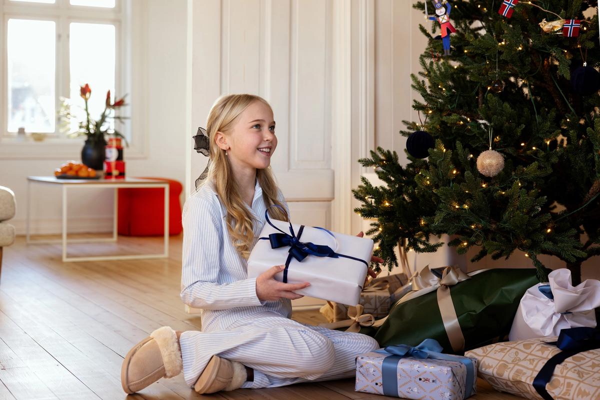Jente med lyst hår i pysj som sitte ved juletreet og holder en pakke i hånden. Smiler til noen utenfor bildet.