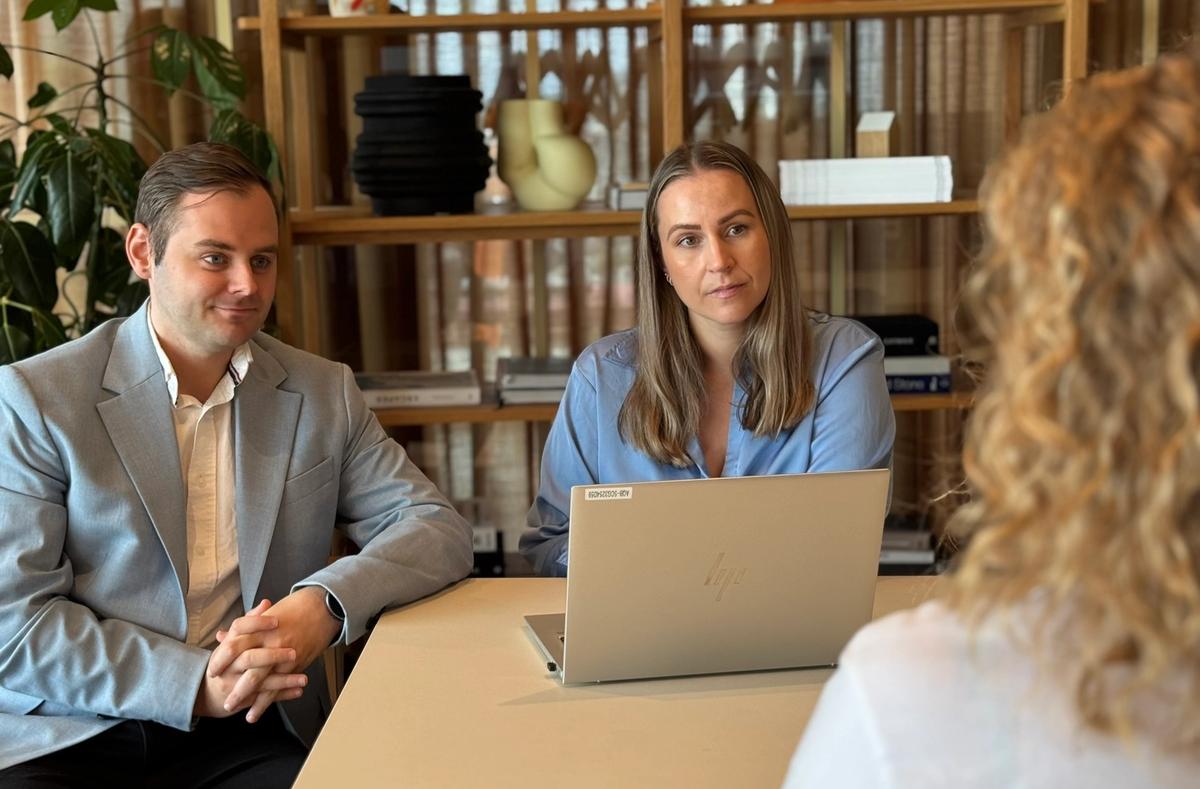 Mann og kvinner som sitter ved en bord med laptop mellom dem og en kvinne. Begge lytter til kvinnen.