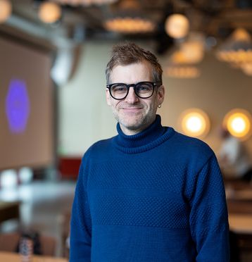 Mann med blå høyhalset genser og briller, smiler og ser i kamera. Blurret bakgrunn.