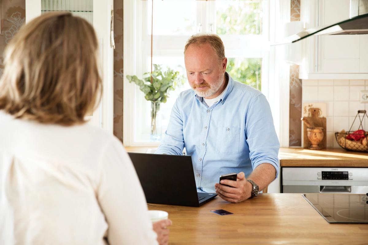 Mann med blå skjorte sitter med pc ved kjøkkenøyen, holde mobil i hånden. Dame sitte med ryggen til.