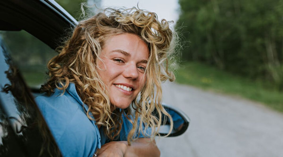 Kvinne lener seg ut av bilvindu, ser inn i kamera og smiler