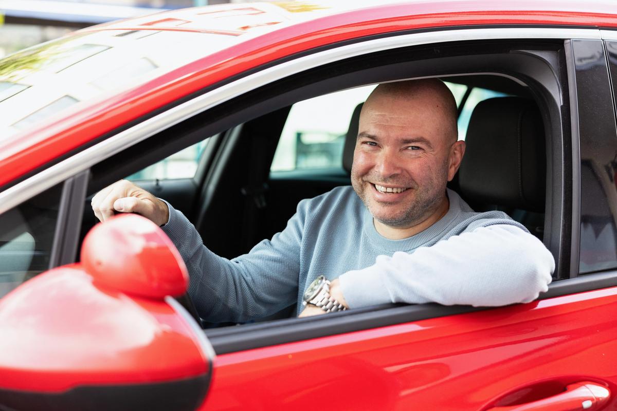 Mann med lys genser i førersetet på en rød bil. En hånd på rattet og den andre på dørkarmen.