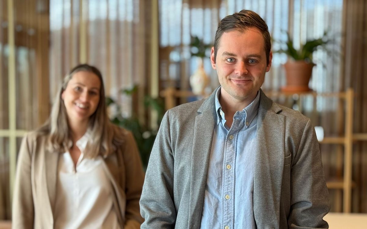 Mann med blå skjorte og grå dressjakke ser inn i kamera og smiler. Til venstre bak mannen, står en kvinne med hvit bluse og beige dressjakke, ser mot kamera og smiler.
