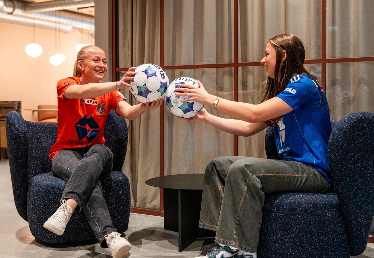 To kvinner med fotballdrakt på sitter i loungestoler og holder fotballer i hendene som de rekker mot hverandre.
