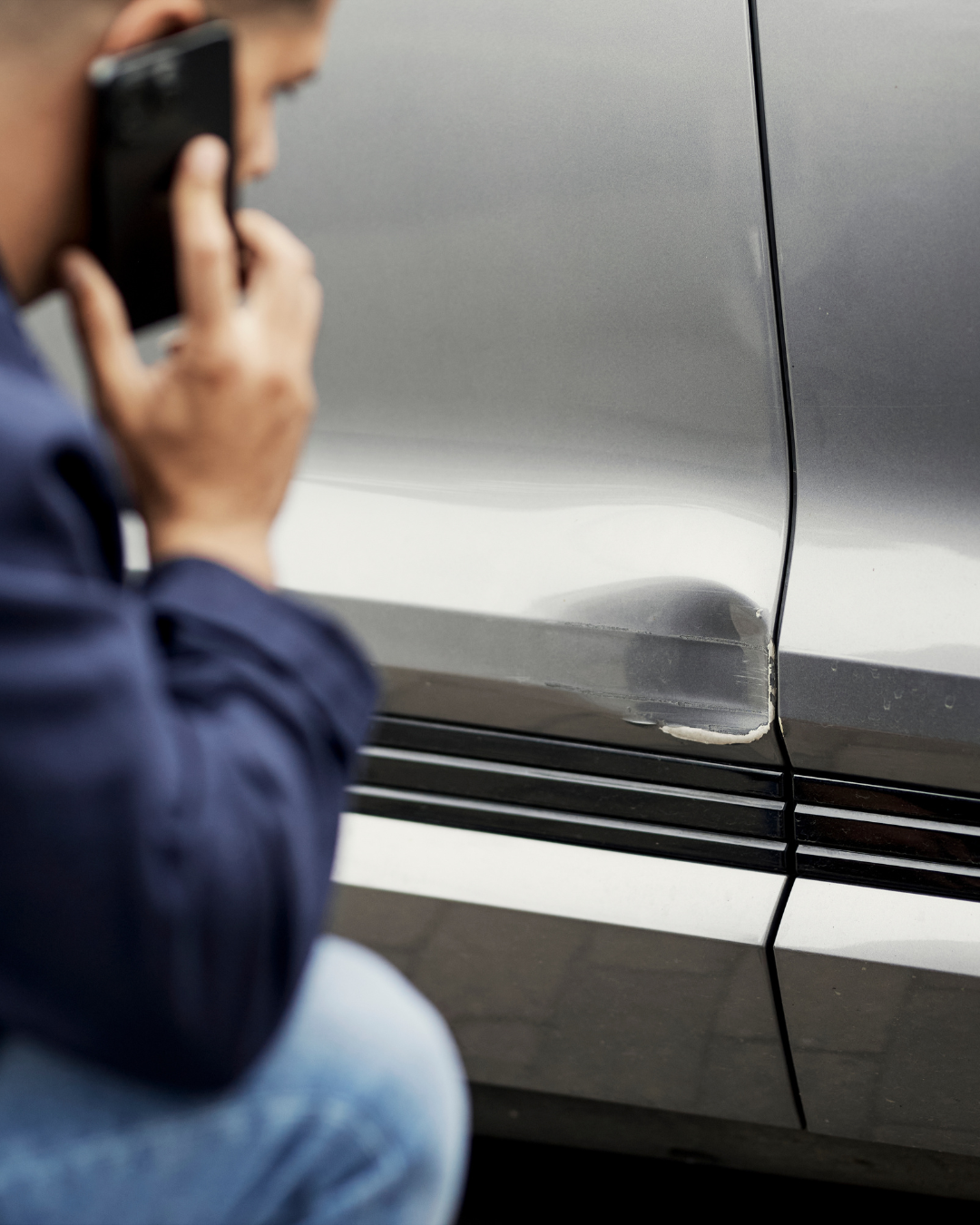 Bildet viser siden på en sølvfarget bil som har fått en bulk. Foran bilen sitter en person på huk.