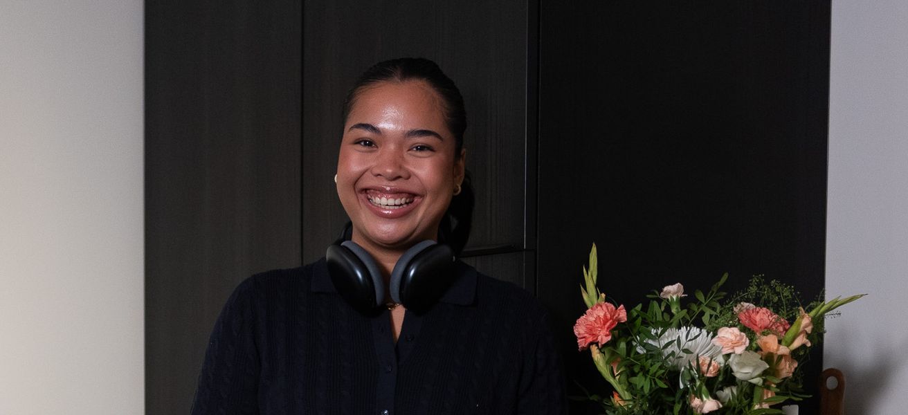 Ung kvinne med headset rundt halsen, på kjøkkenet. Hun står oppreist og smiler til kamera