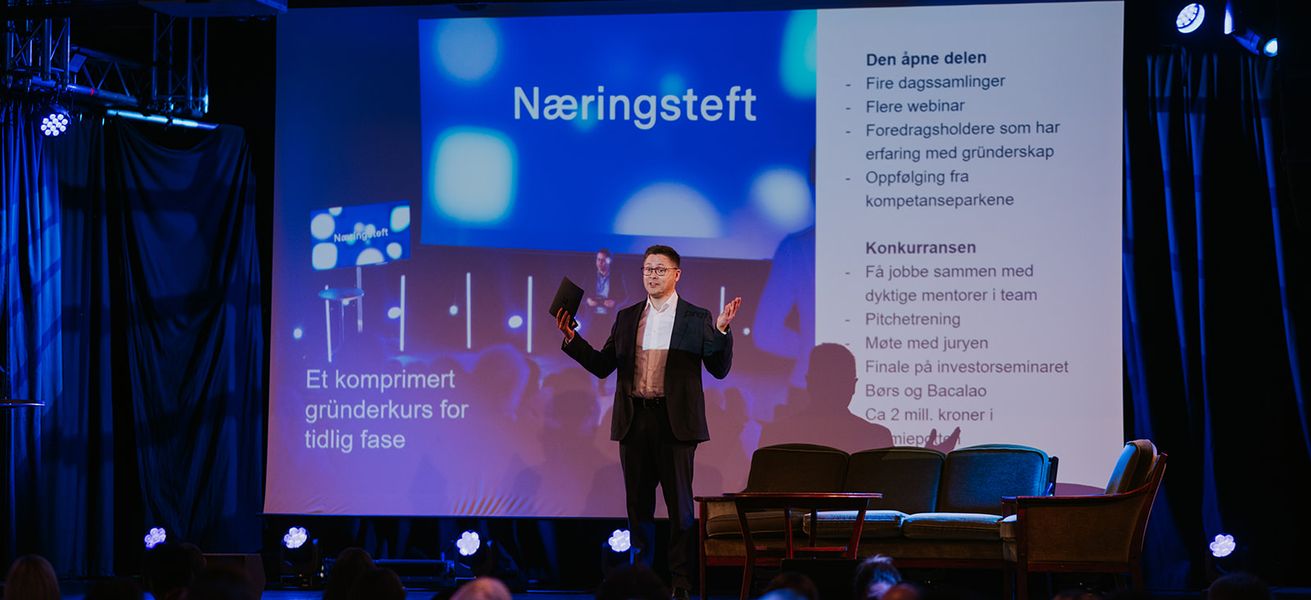 Mann som står på scenen foran et publikum. I bakgrunn vises presentasjon av Næringsteft bak seg.