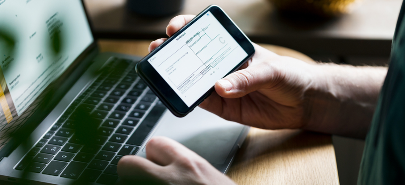 Hender som holder mobiltelefon med en faktura. Laptop i bakgrunn.